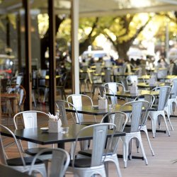 緑豊かな木々に囲まれた、黒いテーブルと金属製の椅子が特徴のカフェの屋外ダイニングエリア。